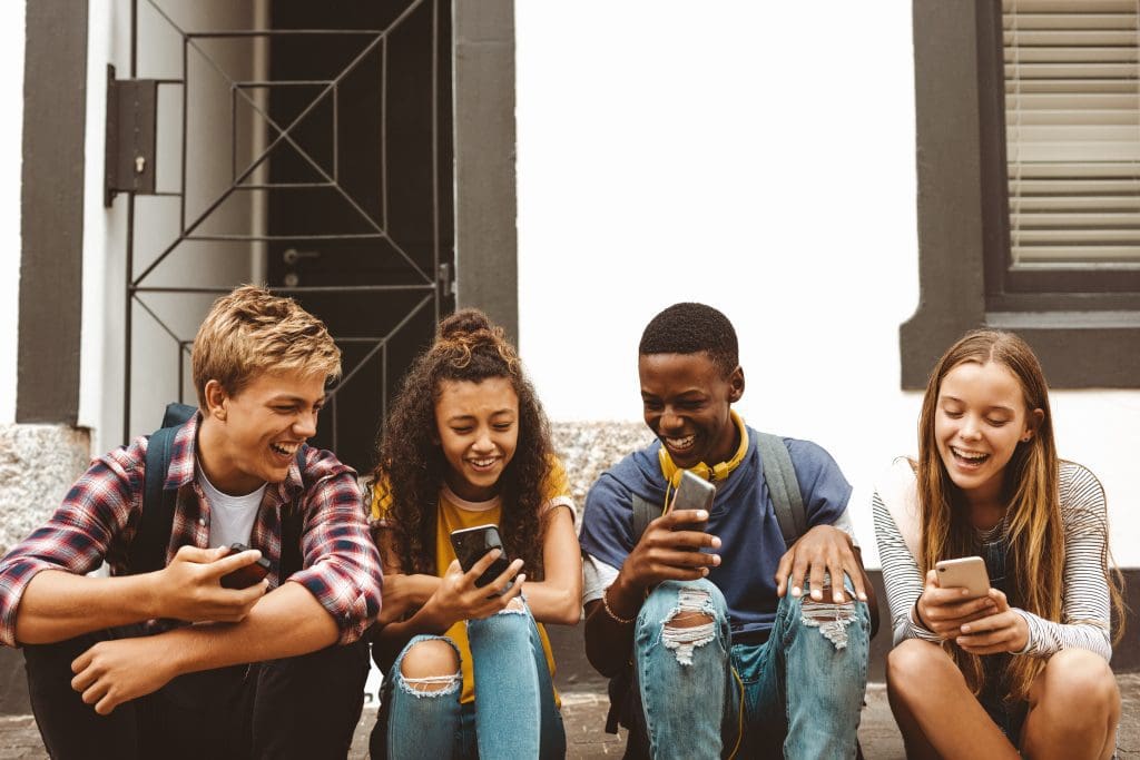 Teenage friends sitting on a pavement holding their mobile phones. Cheerful college boys and girls having fun talking sitting outdoors in a street.