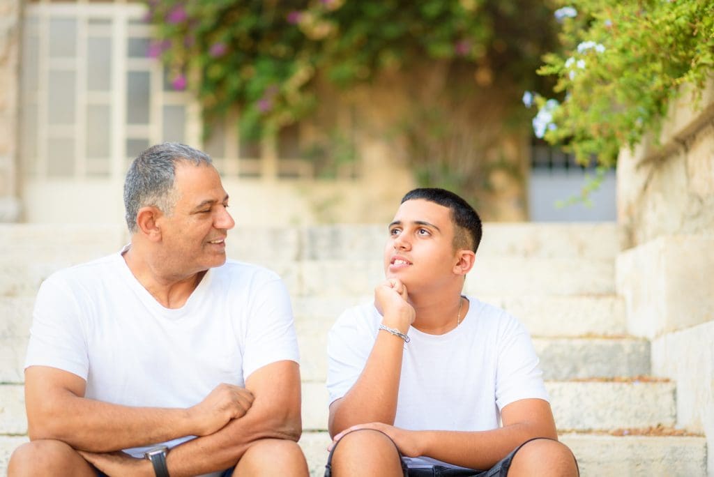 Teenager son and senior father sitting on stairs outdoors at home, talking.