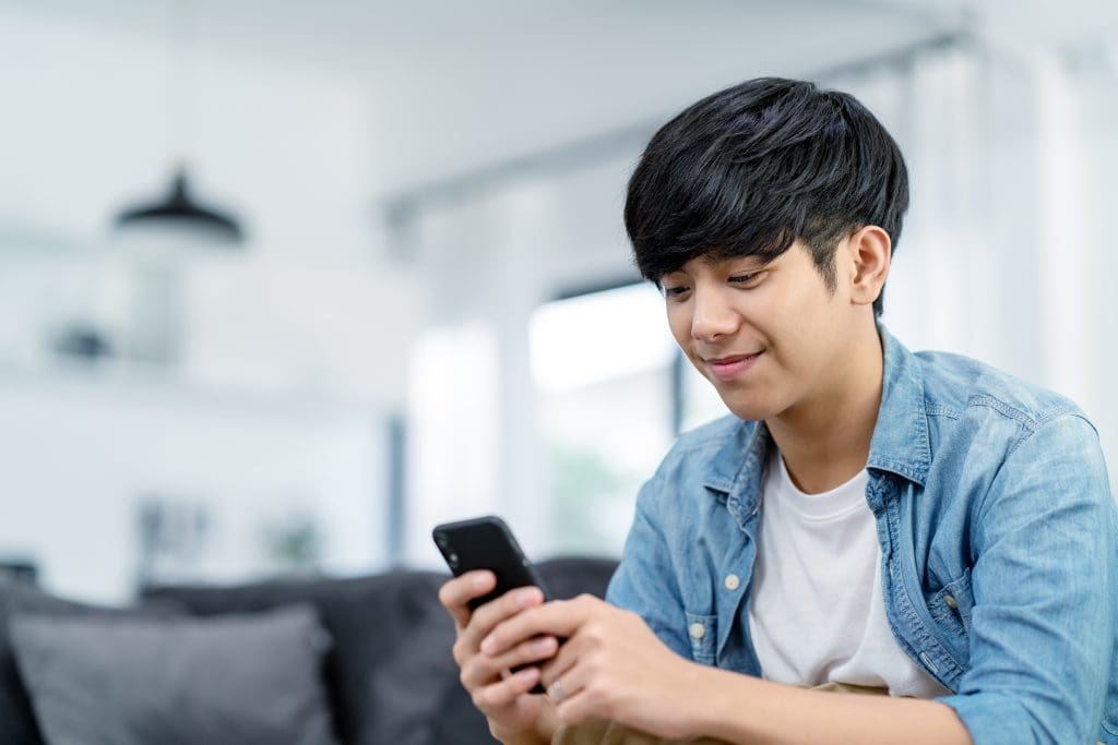 Happy teenager using smart phone and smiling on sofa living room at home. Teen holding and using cellphone for searching data and social media on internet.